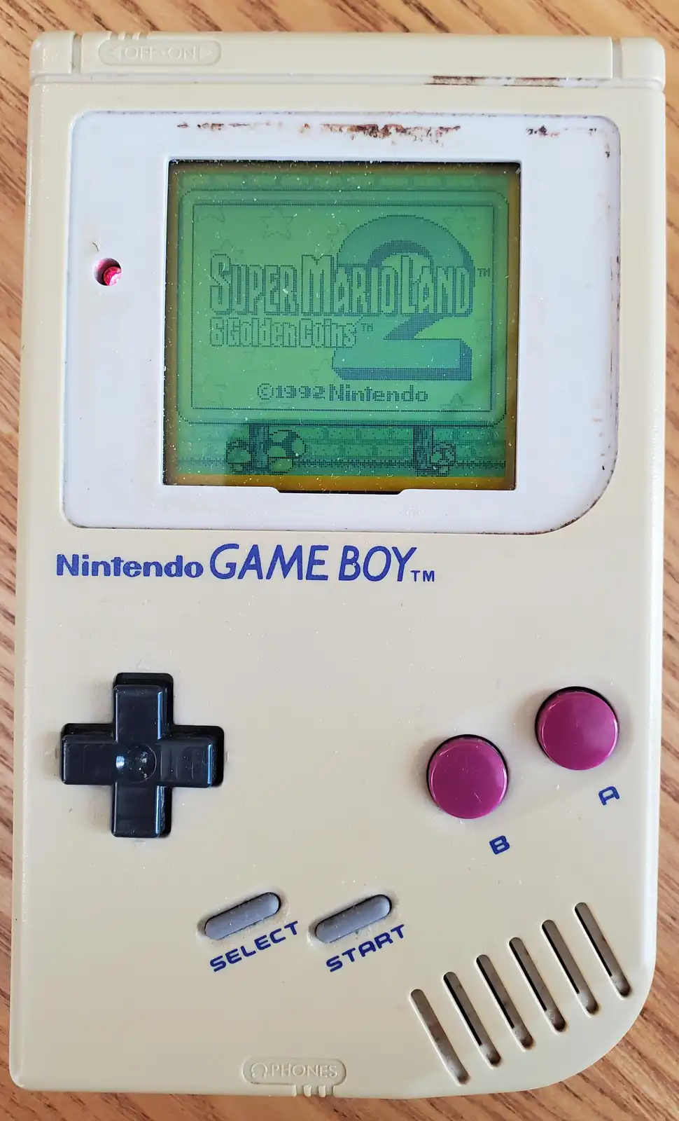 A beige Nintendo Game Boy displays the title screen of 'Super Mario Land 2: 6 Golden Coins' on its green screen, with…