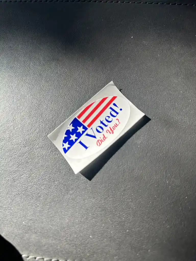 A close-up photo shows a standard American 'I Voted!' sticker, featuring red stripes and white stars on a blue field, stuck…