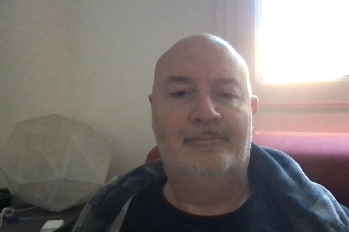 A bald man with a goatee stares blankly at the camera while sitting indoors, with a geometric lamp and window light visible…