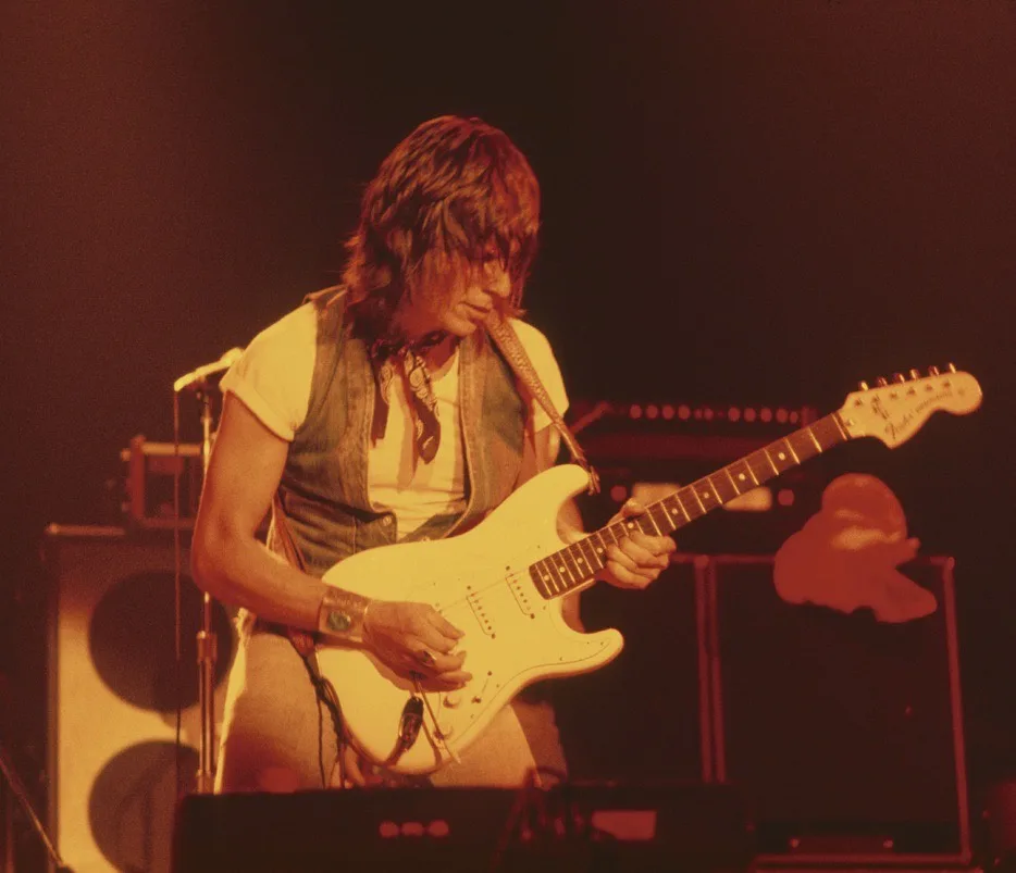 A musician with long hair, wearing a vest over a t-shirt, plays a white electric guitar on stage under warm red lighting…