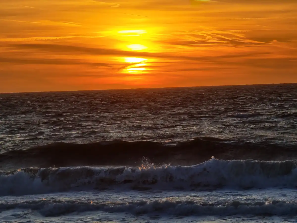 A dramatic sunset over the ocean with fiery orange and yellow clouds, the sun low on the horizon, and dark, choppy waves…