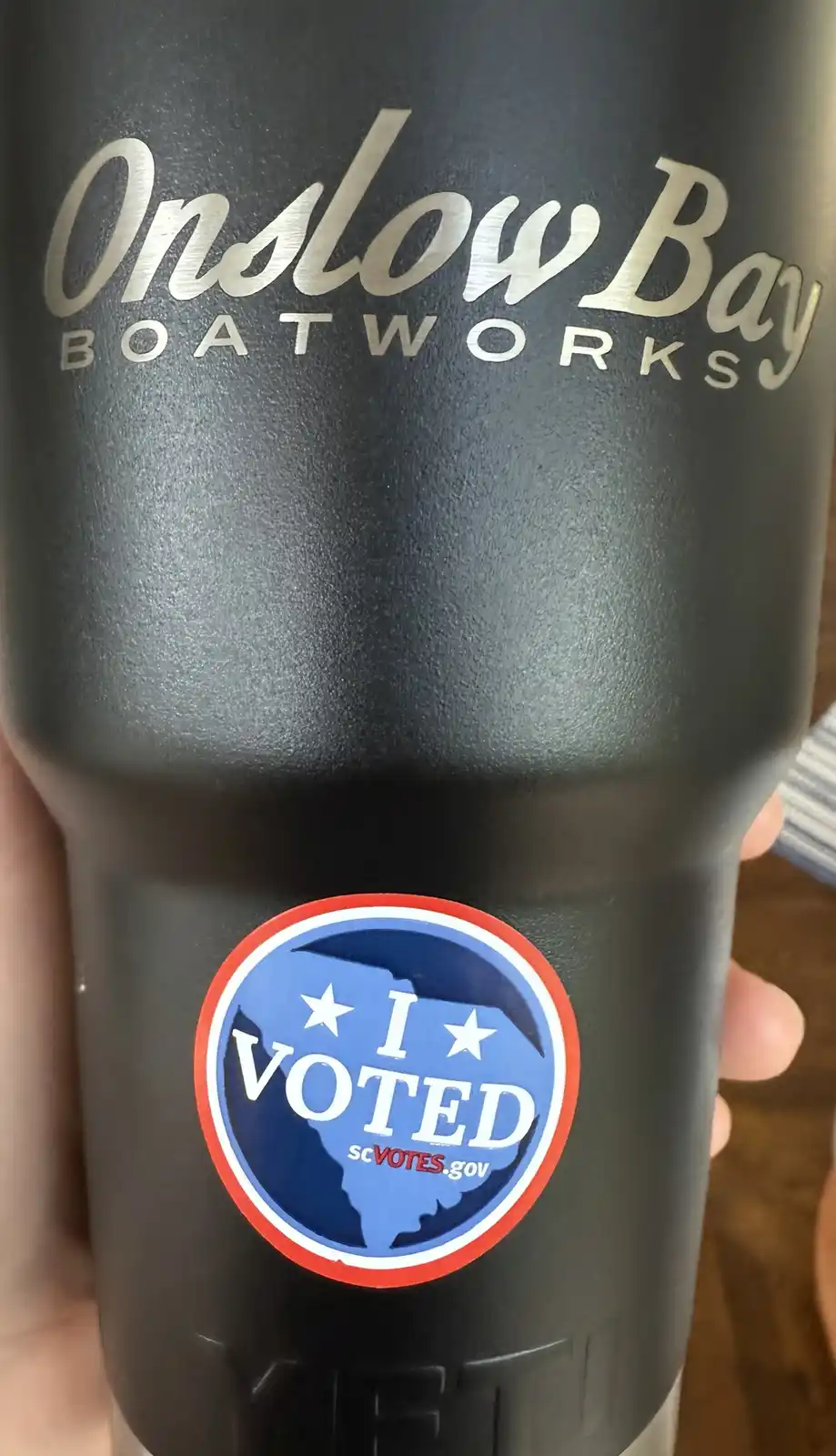 A close-up of a black Onslow Bay Boatworks branded tumbler held in a hand, featuring a circular 'I VOTED' sticker with a…