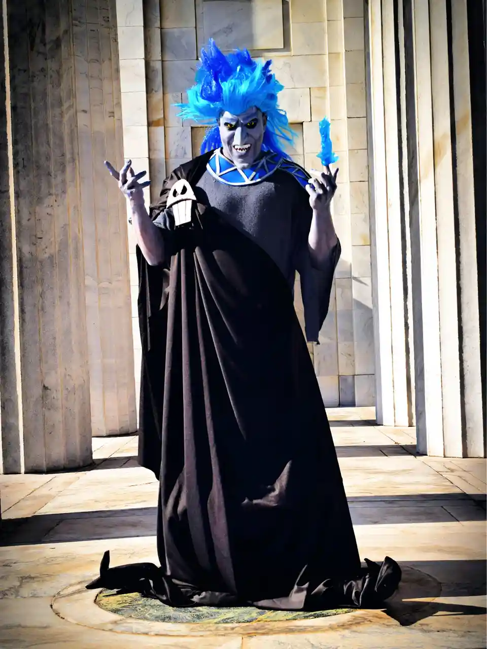 A person in a dramatic costume with blue spiky wig, grey face paint, and black robe stands in front of marble columns…