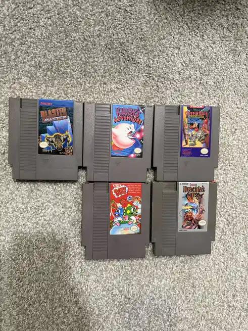 Five gray Nintendo Entertainment System game cartridges are laid out on a gray carpet. From left to right, top row: Blaster…