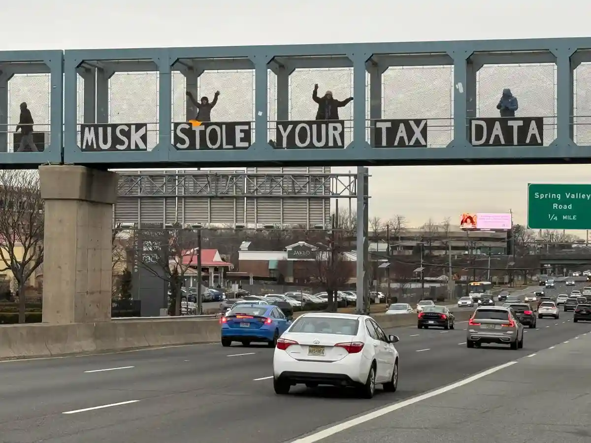 People stand on a pedestrian overpass above a highway, each positioned behind a section of a large sign that collectively…