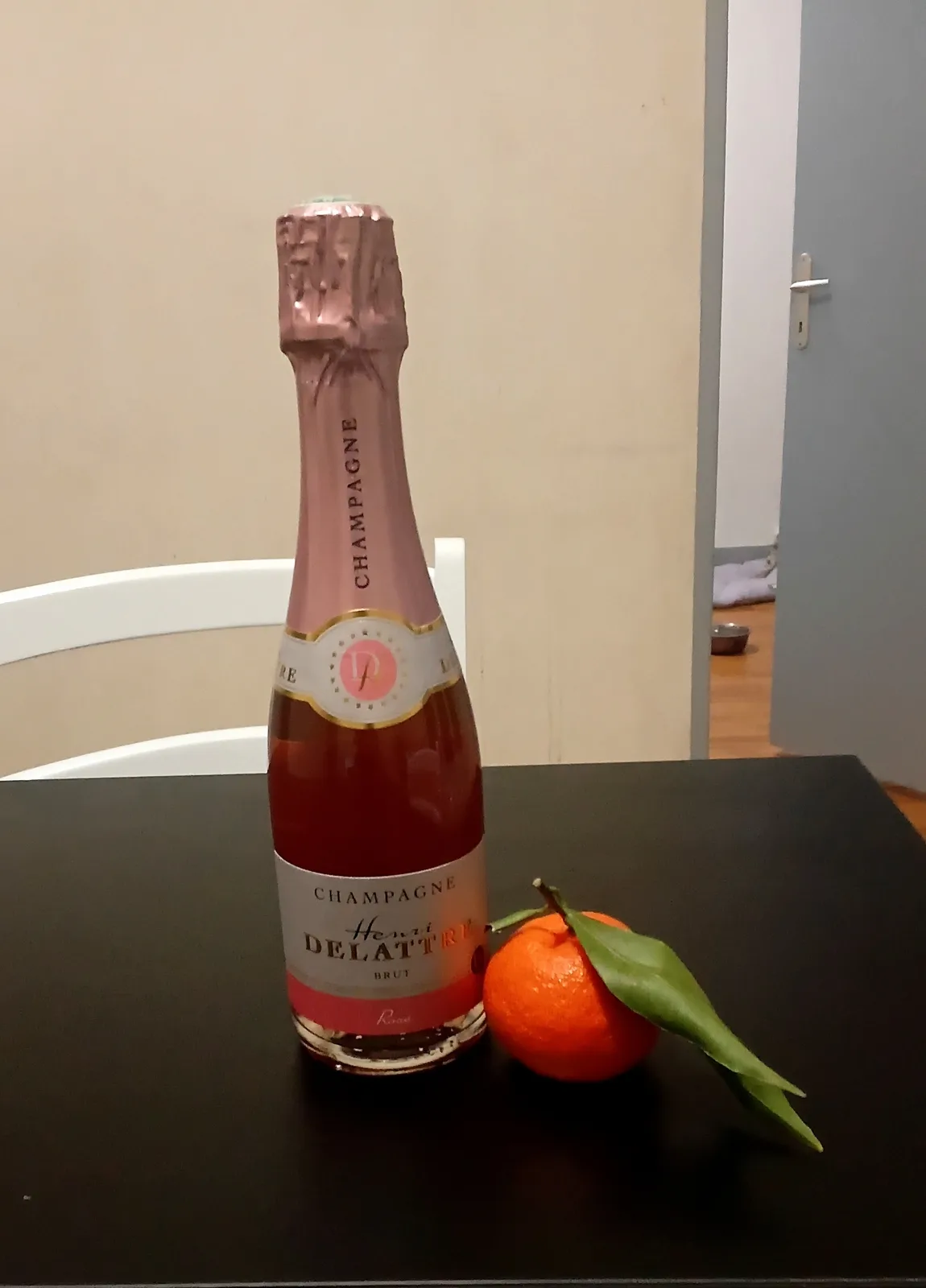 A bottle of pink champagne labeled 'Henri Delattre Brut' sits on a dark table next to a small orange tangerine with a green…
