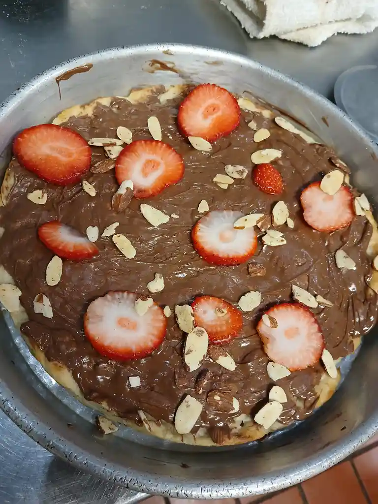 A round pizza in a metal pan is covered with a thick layer of chocolate spread, topped with sliced strawberries and sliced…