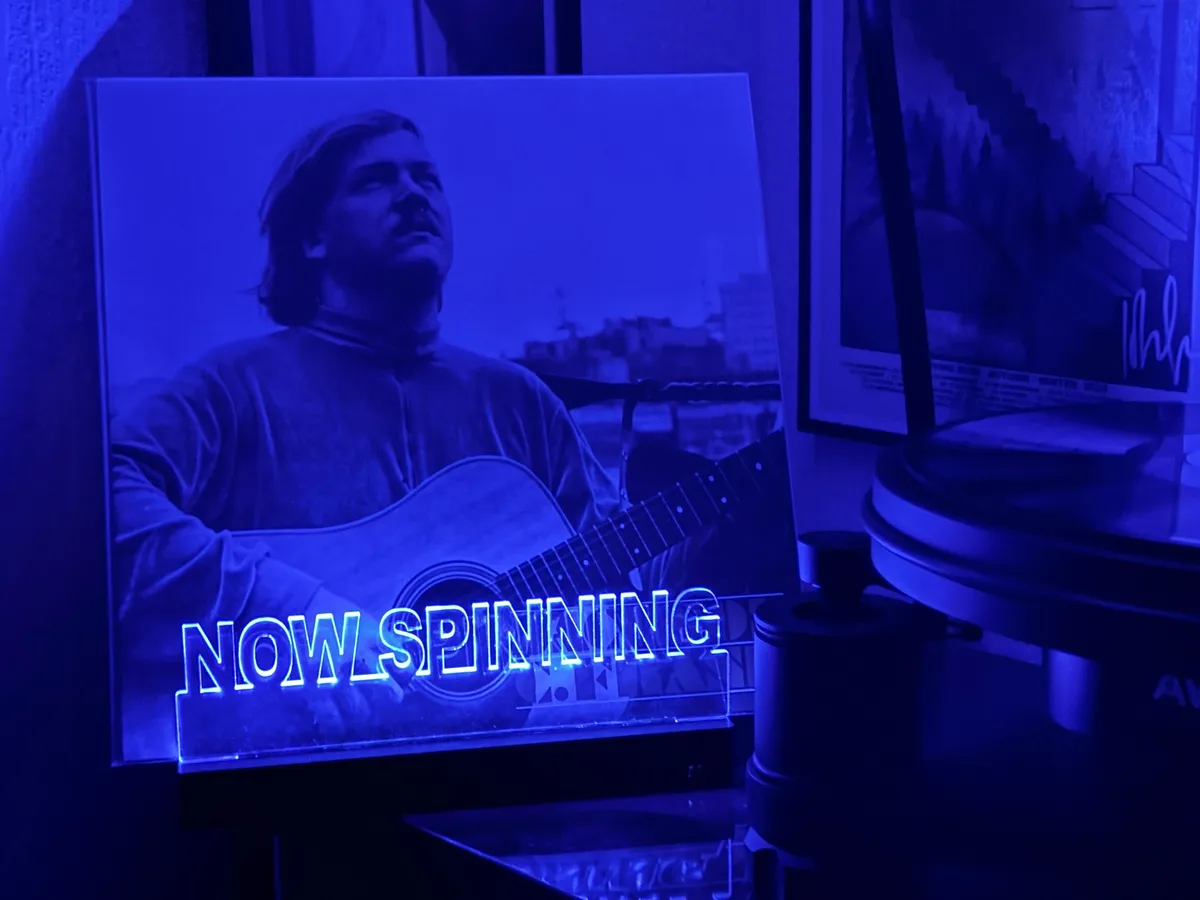 A blue-lit photo of a man with a guitar is displayed on a stand next to a record player, with glowing neon text reading 'NOW…