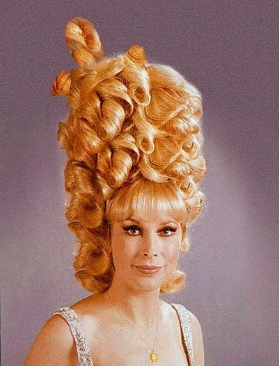 a woman with a very large, elaborate blonde beehive hairstyle made of tight curls, wearing a sparkly top and a necklace…