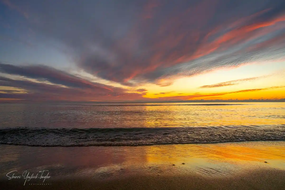 A serene beach sunset photo with vibrant orange, pink, and purple clouds reflecting on calm ocean water and wet sand. The…