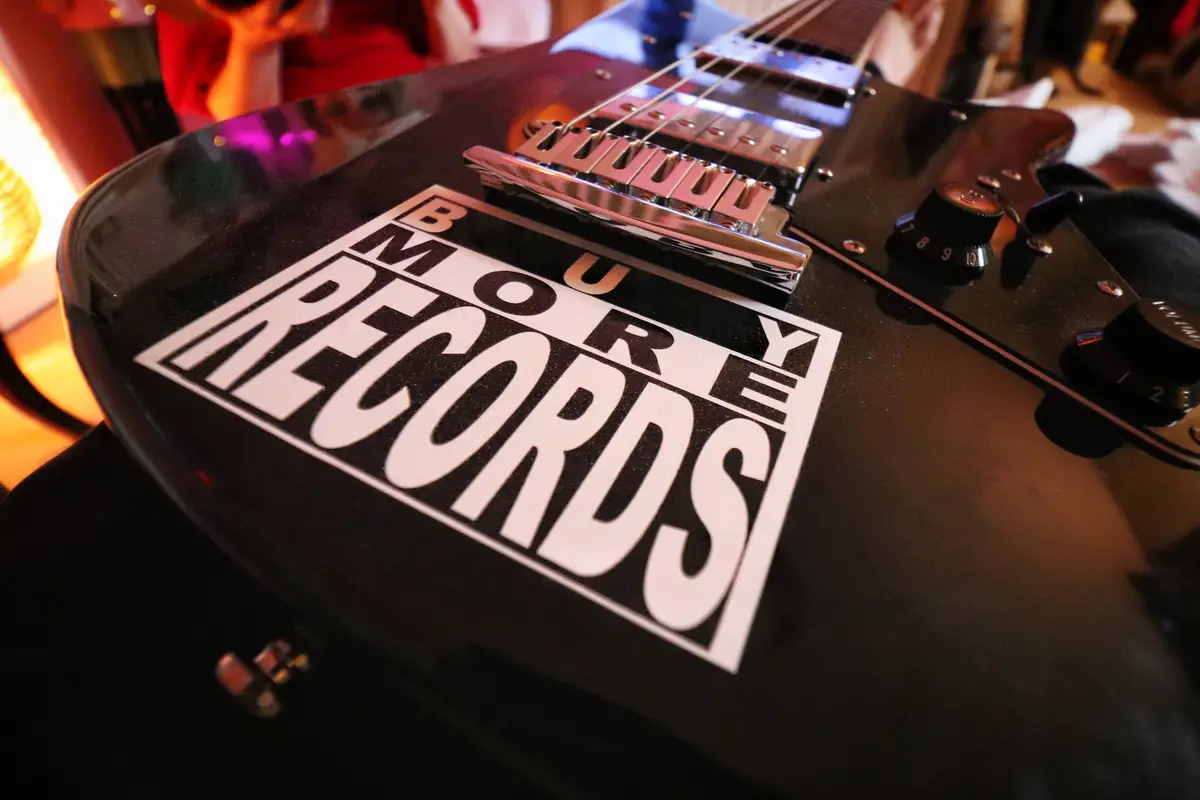 A close-up shot of a black electric guitar with a white sticker that reads 'MORE RECORDS' in bold, stylized lettering. The…
