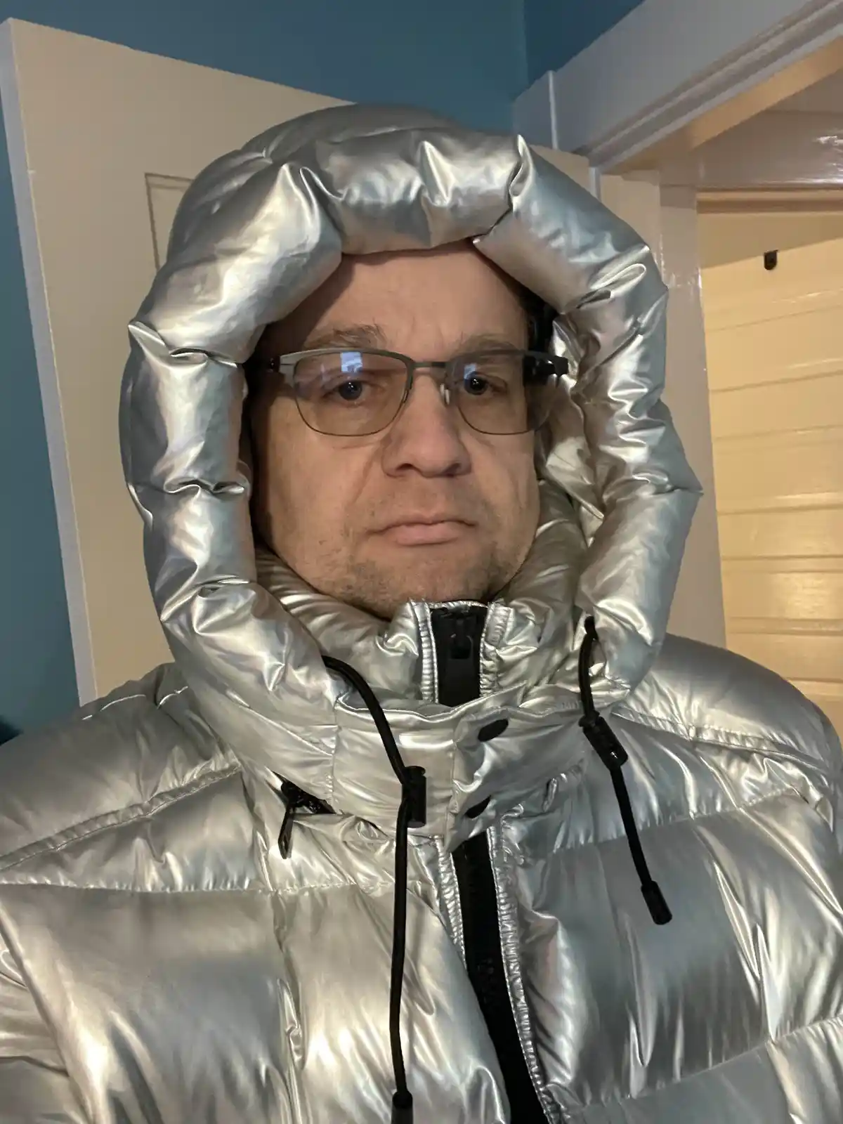 A man with glasses wears a shiny silver puffer jacket with the hood pulled up over his head, resembling a helmet. The text…