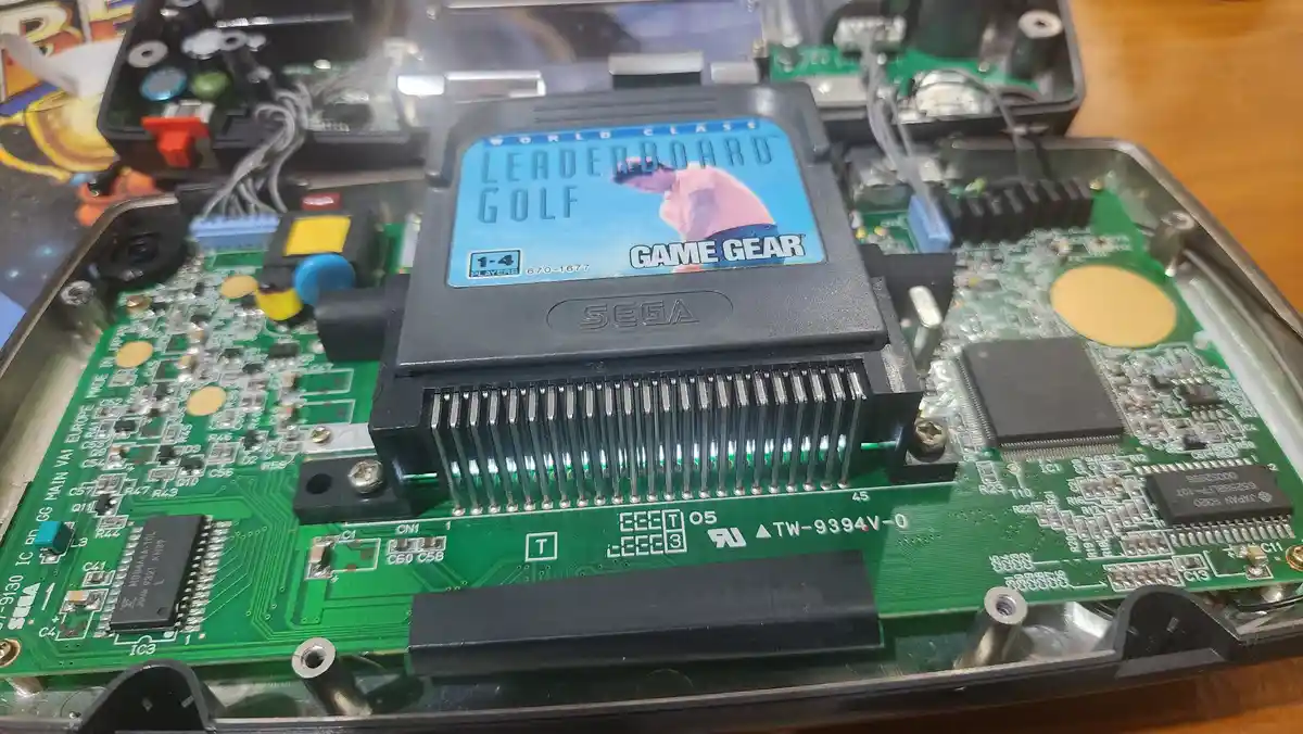A close-up photo of a Sega Game Gear handheld console with its back panel removed, revealing the green circuit board and…