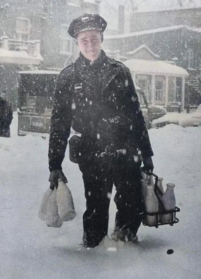 A vintage black-and-white photo shows a smiling milkman in a uniform walking through deep snow, carrying milk bottles in one…