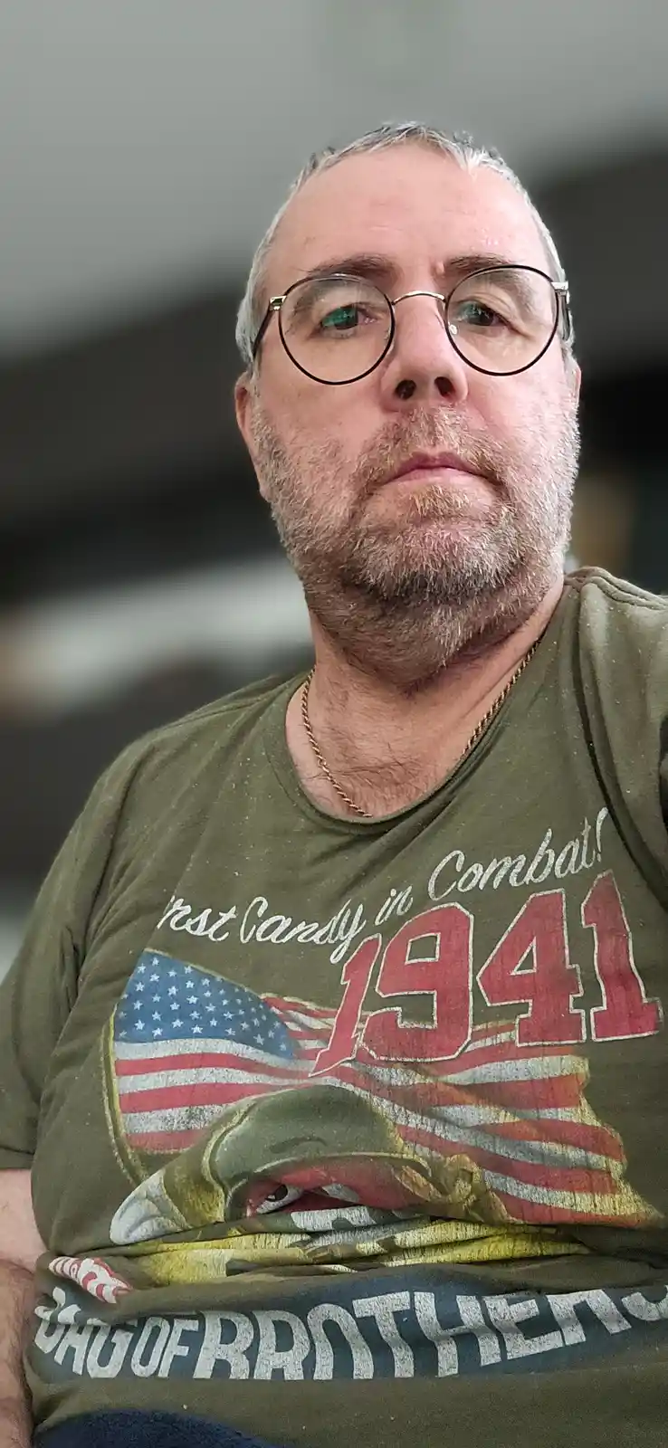 A middle-aged man with glasses and a graying beard wears an olive green t-shirt featuring a graphic of an American flag and…