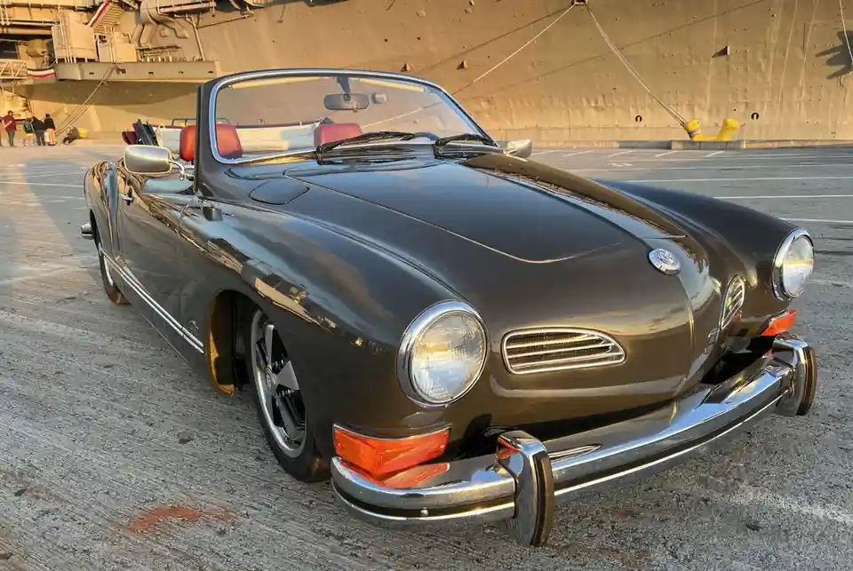 A dark brown vintage Volkswagen Karmann Ghia convertible is parked on a concrete surface with a large ship in the…