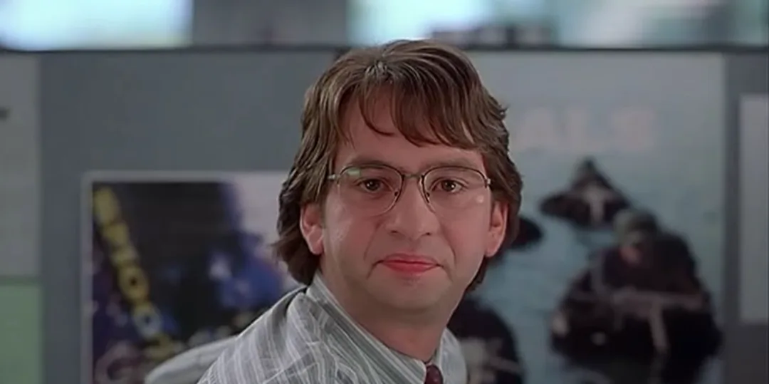 a man with brown hair and glasses stares directly at the camera with a subtle, slightly smug expression. He wears a striped…