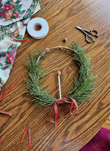 An overhead shot shows a partially assembled Christmas wreath made of pine needles on a wooden table, surrounded by crafting…