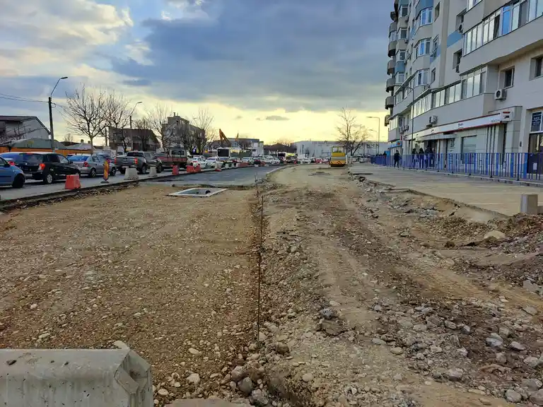 A street is under construction with a large, uneven dirt and gravel patch in the middle, flanked by a paved road with cars…