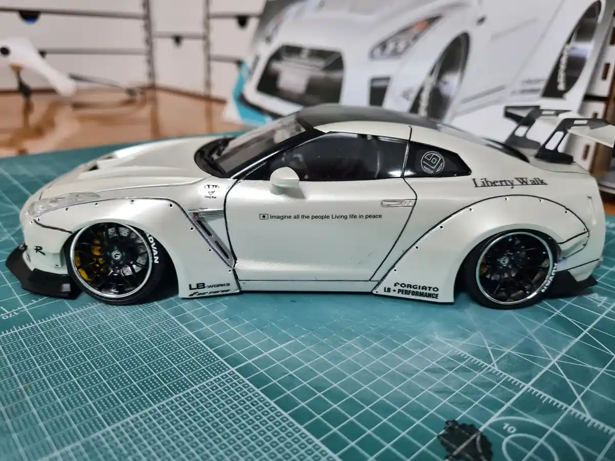A white scale model of a modified Nissan GT-R with widebody kit and black wheels sits on a green cutting mat. The car has…