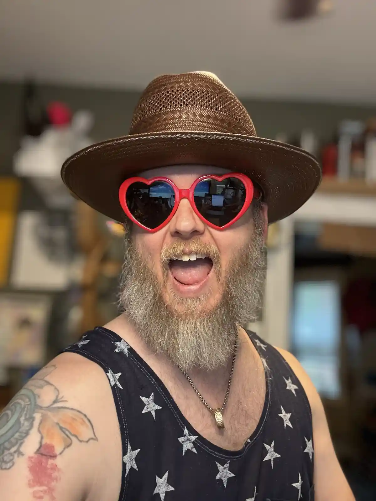 A man with a full gray beard and mustache wears a brown straw hat and oversized red heart-shaped sunglasses while making a…