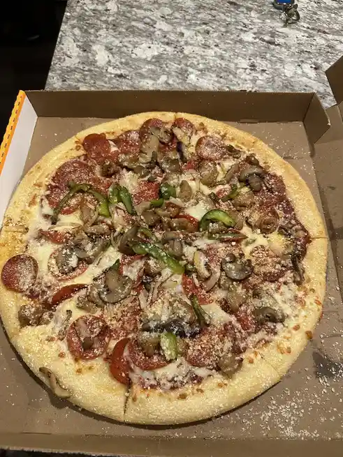 A close-up photo of a pepperoni, mushroom, and green pepper pizza in an open cardboard box on a granite countertop. The…