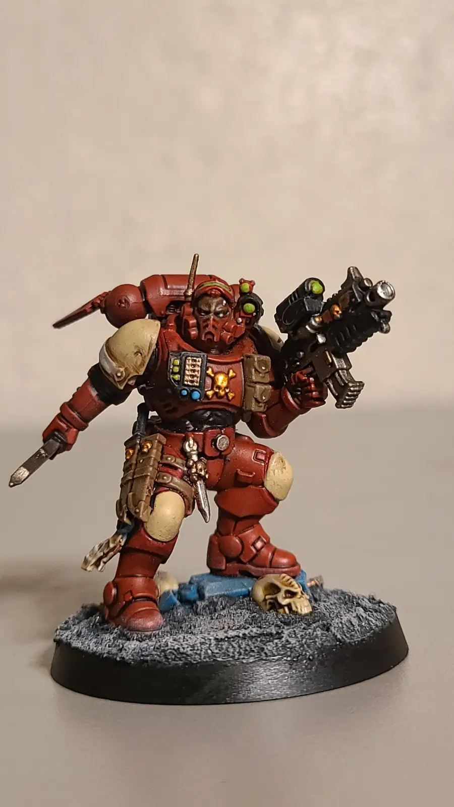 A painted miniature of a red-armored Space Marine, likely from Warhammer 40,000, stands on a grey, rocky base with a skull…