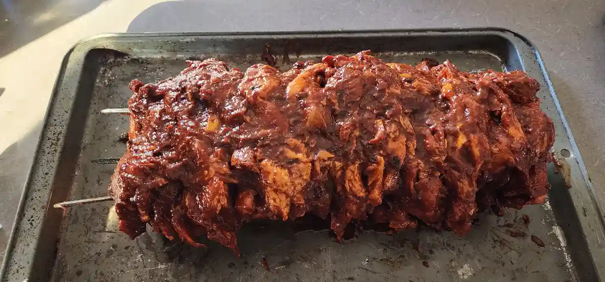 A large, glistening, saucy meatloaf-like dish sits on a metal baking tray, skewered with metal rods. The text overlaid on…