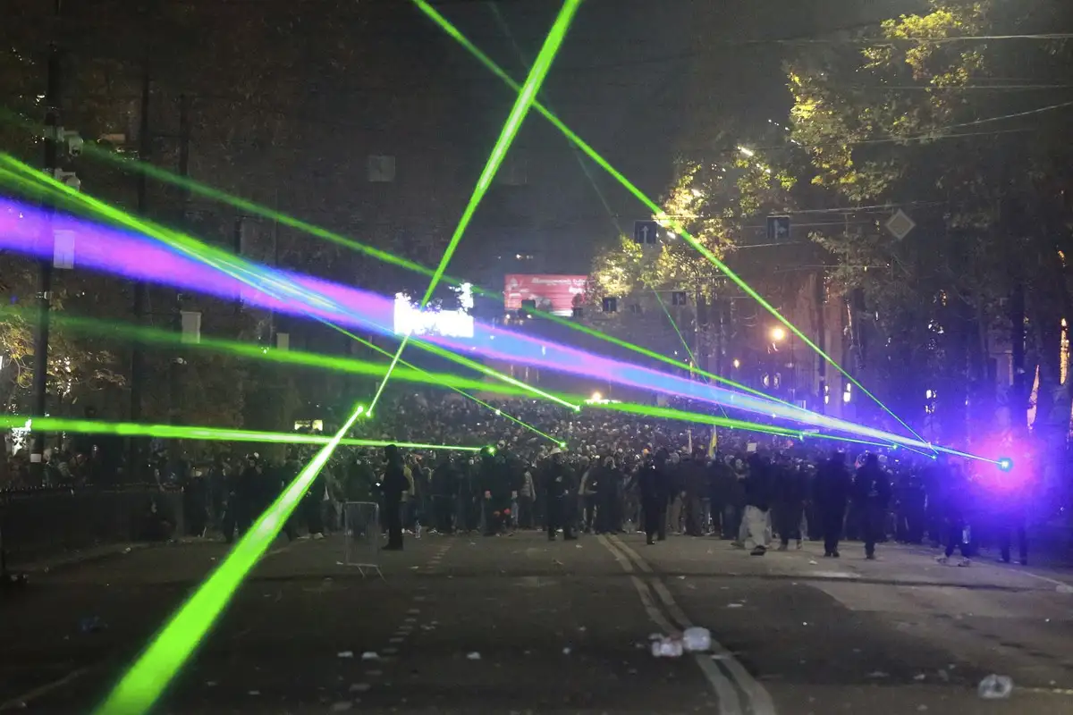A nighttime street scene with a large crowd of people, illuminated by numerous intersecting green and purple laser beams…