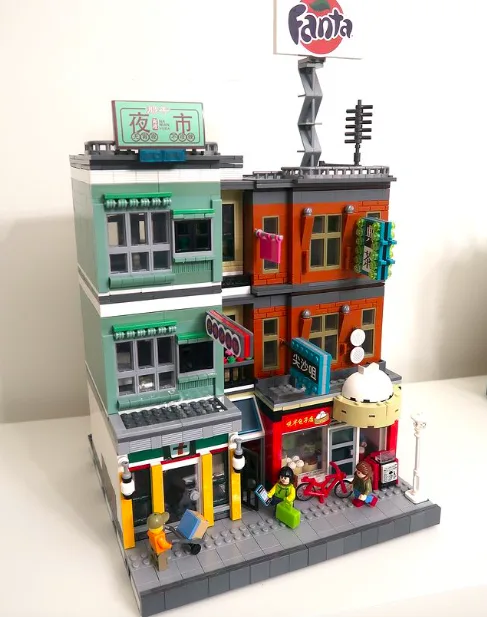 A detailed LEGO model of a multi-story street corner with Chinese characters on signs, including '夜市' (night market) and…