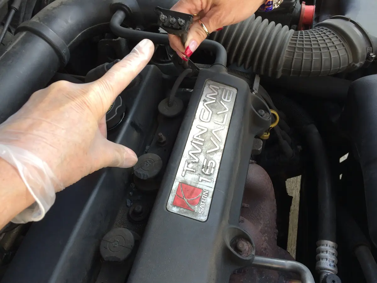 A close-up of a car engine bay shows a hand in a clear plastic glove pointing at a component while another hand with red…