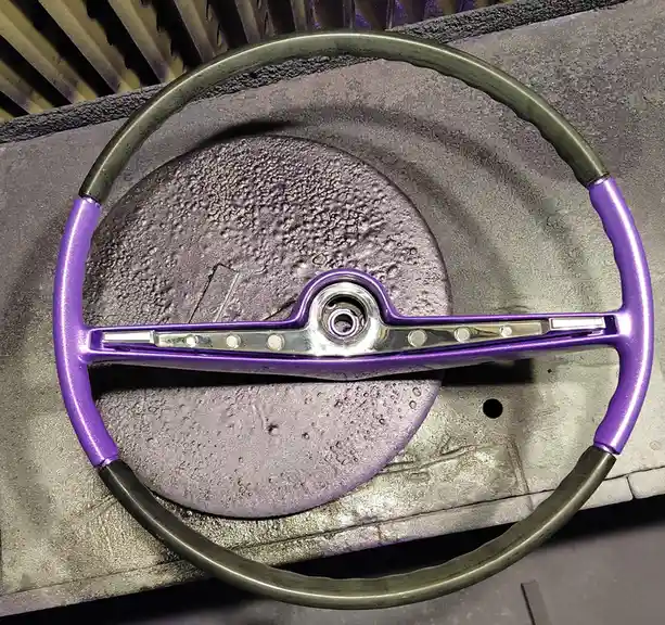 A close-up photo of a vintage-style car steering wheel with a two-tone design: purple on the spokes and handlebars, and a…