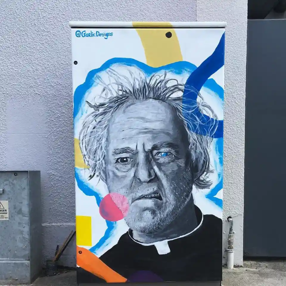 A painted mural on a utility box depicts a man with wild gray hair, a stern expression, and one blue eye, wearing a black…