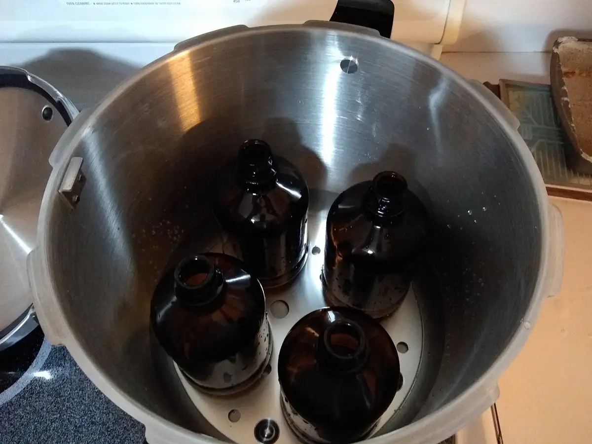 A stainless steel pressure cooker sits open on a kitchen counter, containing four dark amber glass bottles with their tops…
