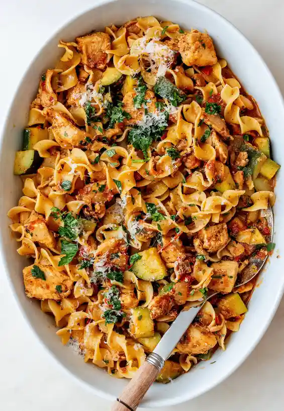 A close-up photo shows a white oval bowl filled with pasta, chicken, zucchini, and herbs, garnished with grated cheese. A…