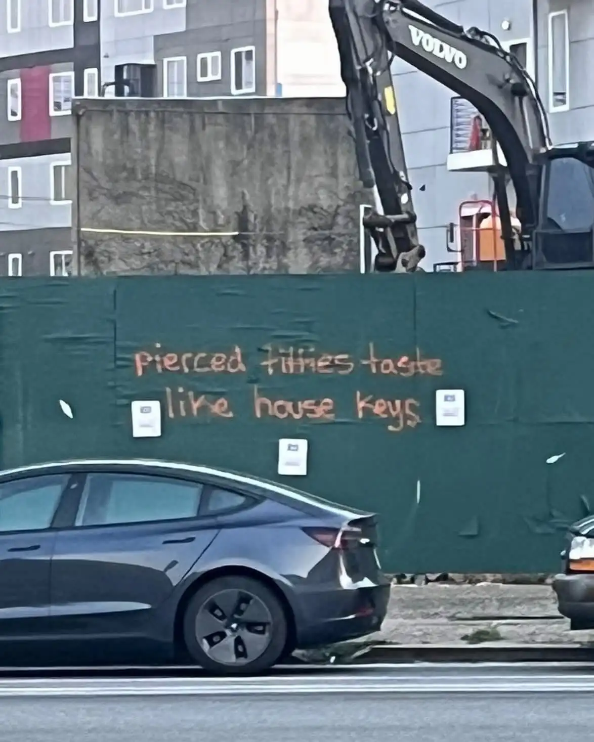 A dark grey Tesla Model 3 drives past a green construction fence where orange spray-painted text reads 'pierced fillies…