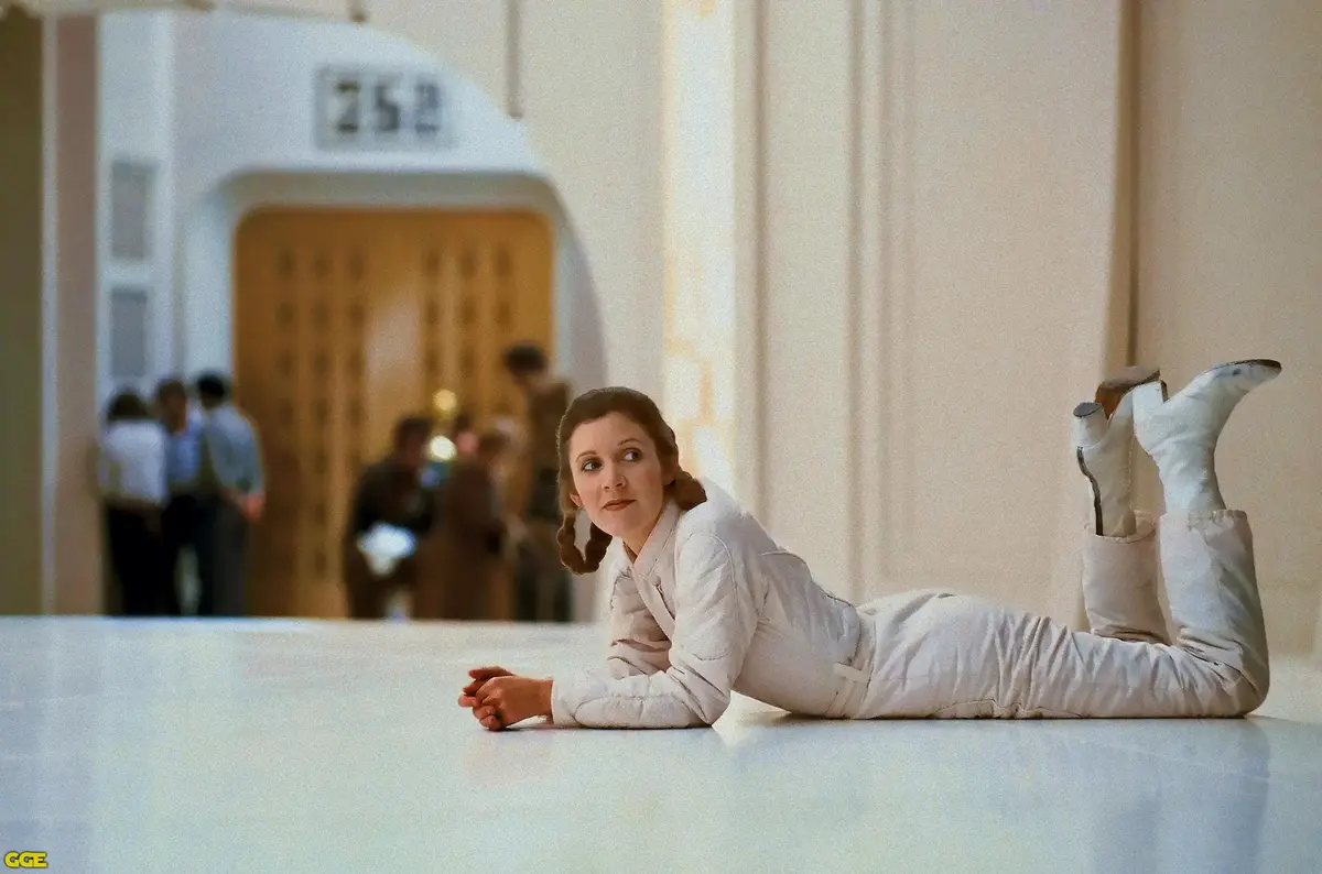 A woman in a white outfit lies on a white floor with her legs kicked up, looking off to the side. In the background, a door…