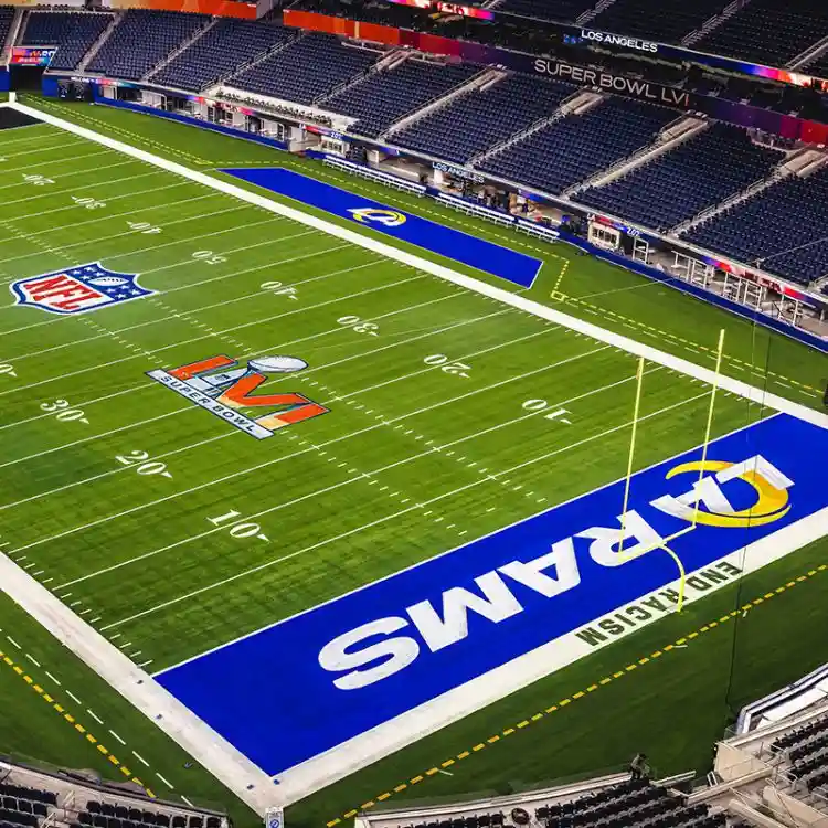 An aerial view of an empty football stadium with a green field and blue end zone. The end zone prominently displays the word…