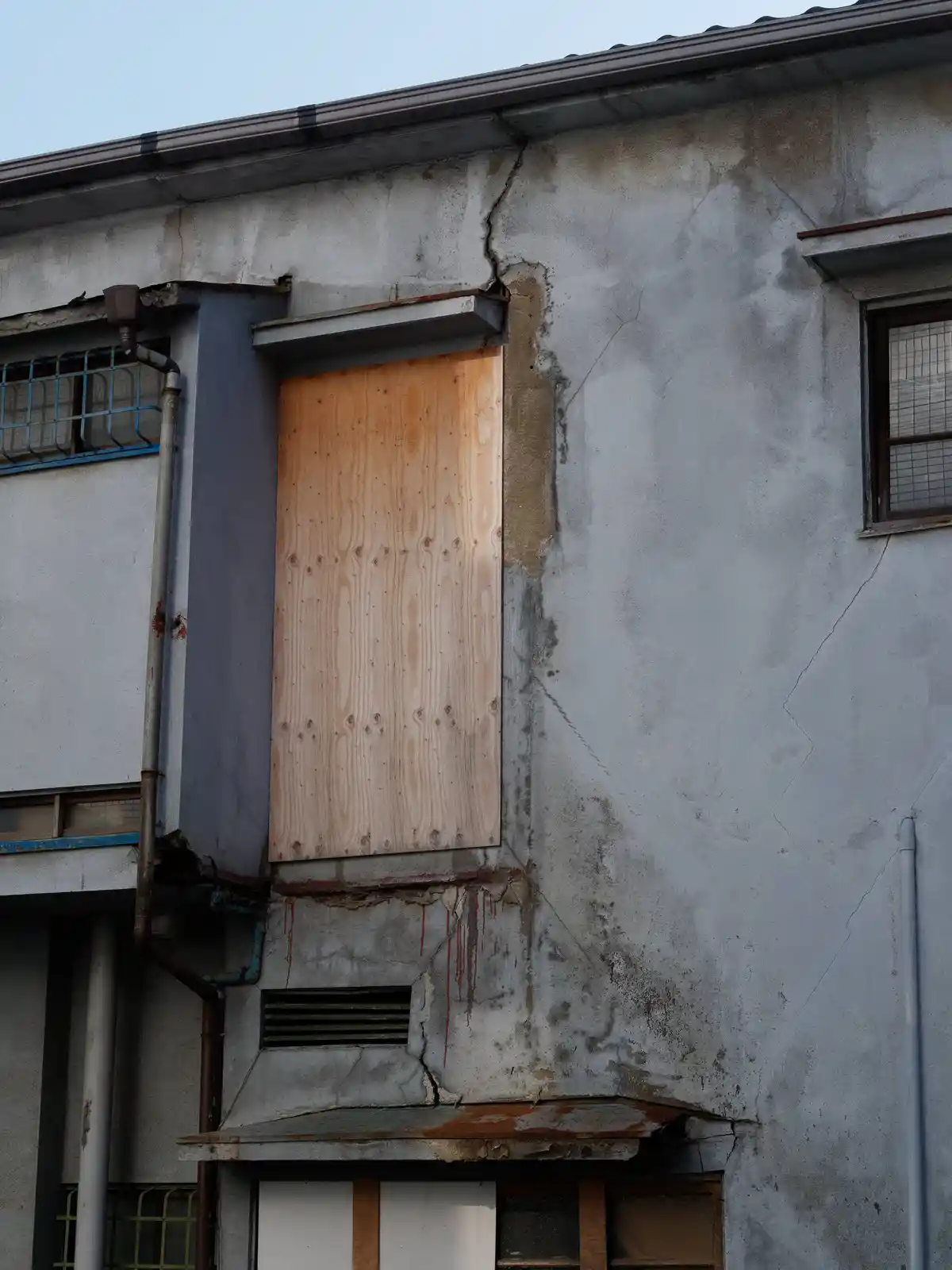 The image shows a weathered concrete building with visible cracks and rust stains. A plain plywood board covers a window…