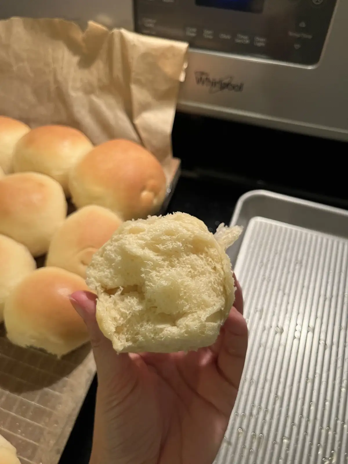 A hand holds up a torn-open, fluffy, golden-brown bread roll, revealing its soft, airy interior. In the background, a baking…