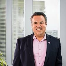 A man in a dark suit and pink shirt smiles broadly while standing in front of a window with a city view. The image has text…