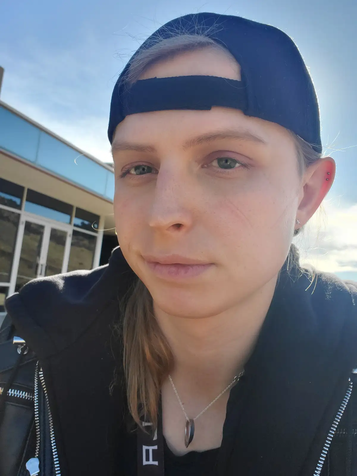 A person with light brown hair wearing a black baseball cap backwards and a black jacket looks directly at the camera with a…