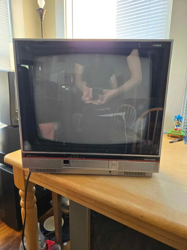 A person holding a phone is reflected in the screen of an old CRT television sitting on a wooden table. The TV is off…
