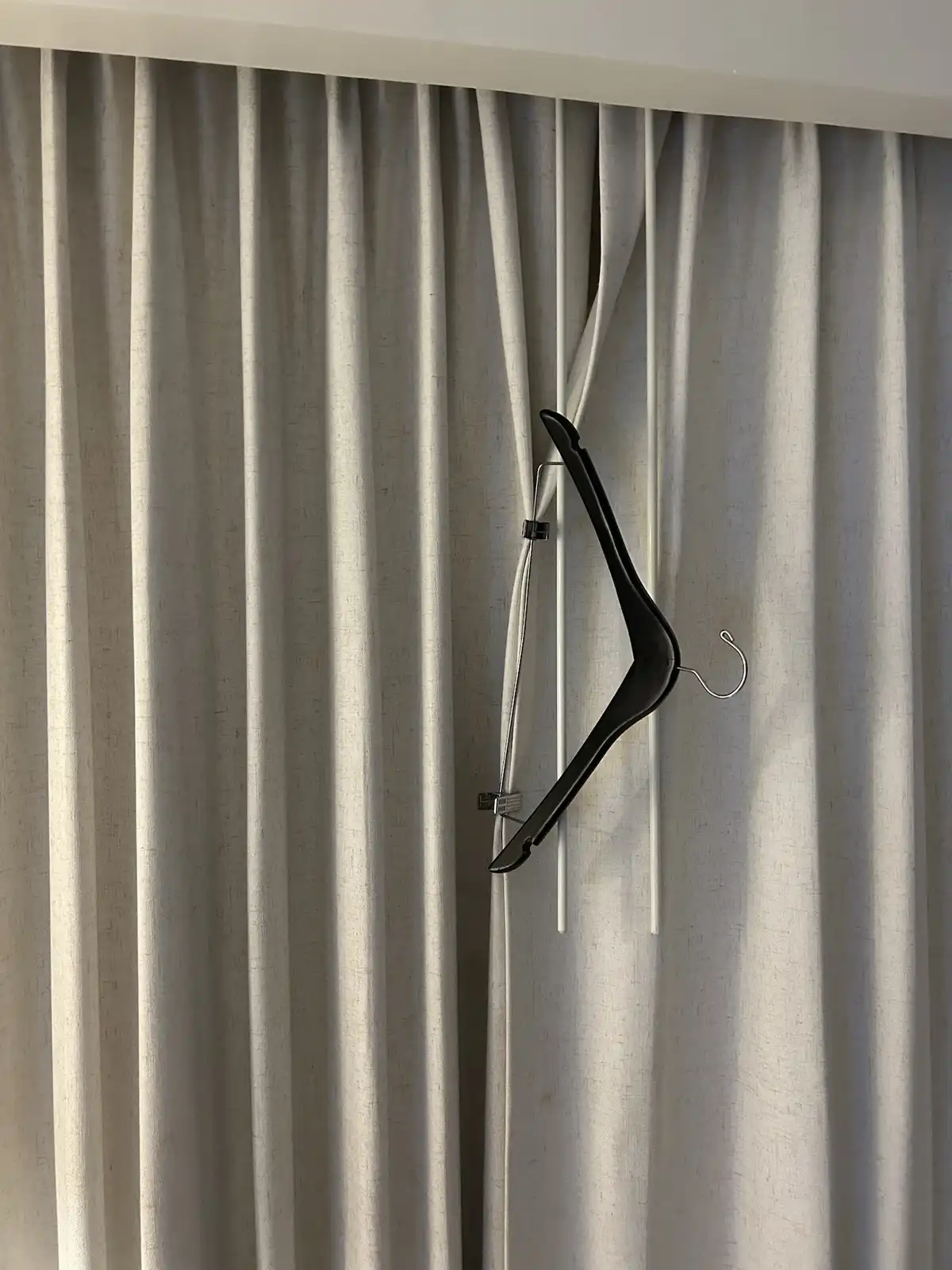 A black plastic clothes hanger is clipped onto the white drawstring cords of light gray curtains, appearing to hang in…