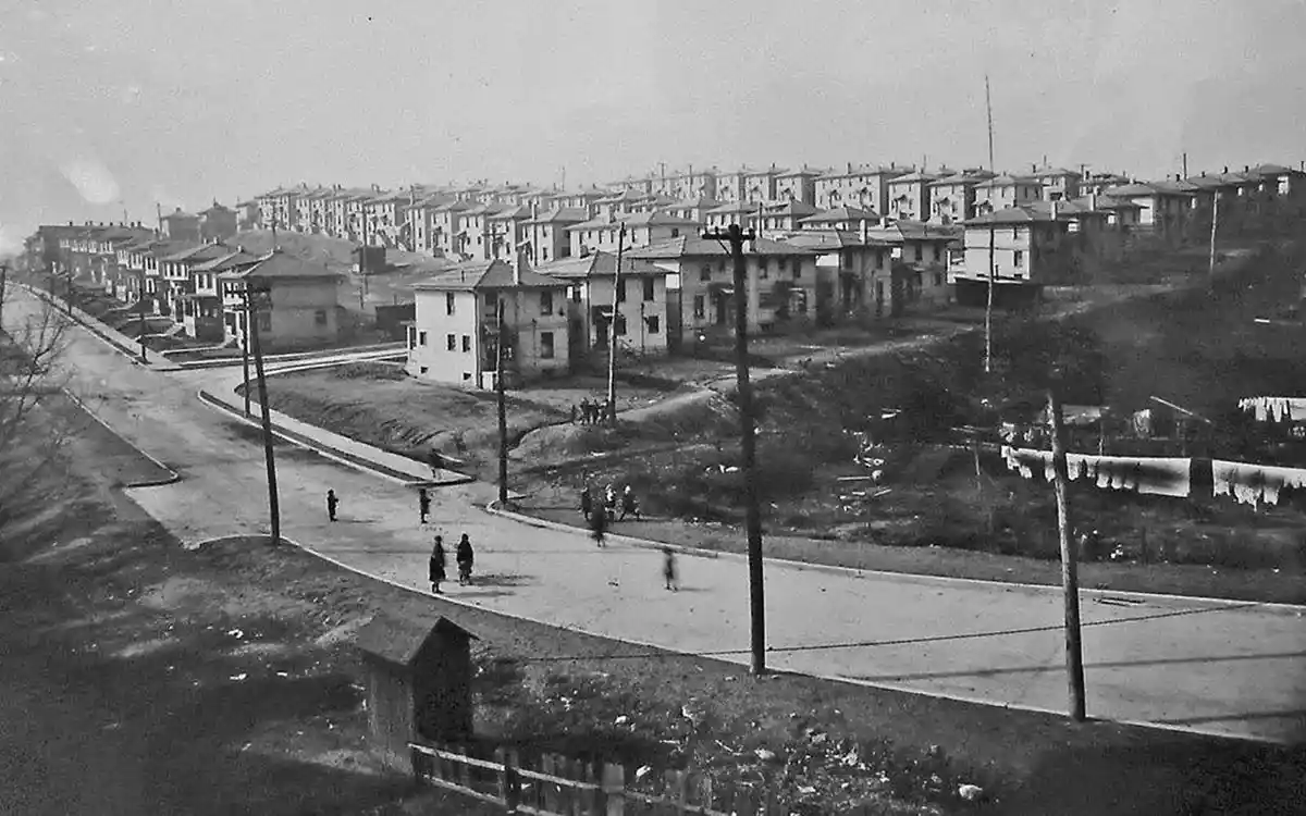 A black-and-white historical photograph shows a wide, curving road with sparse pedestrians, flanked by rows of identical…