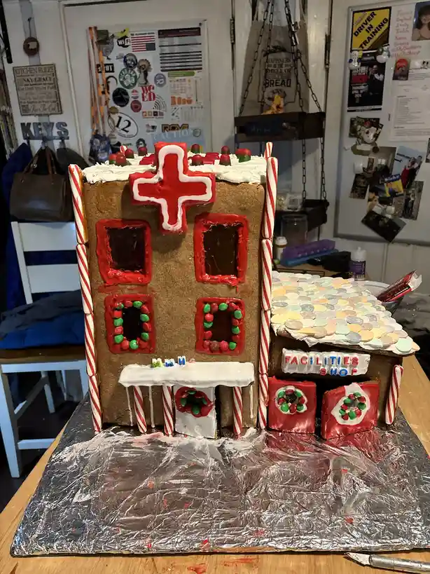 A gingerbread house decorated with candy canes, red icing, and M&Ms, featuring a large red cross on the roof and a sign that…