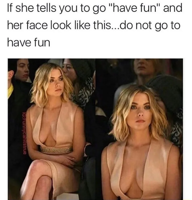 The image shows a woman with blonde hair wearing a revealing beige dress, seated in what appears to be a dimly lit public…