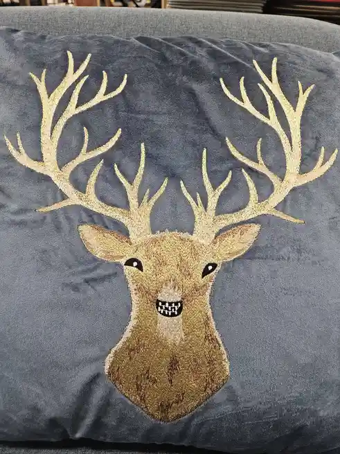 A close-up of a dark blue velvet pillow featuring an embroidered deer head with large, intricate golden antlers. The deer…