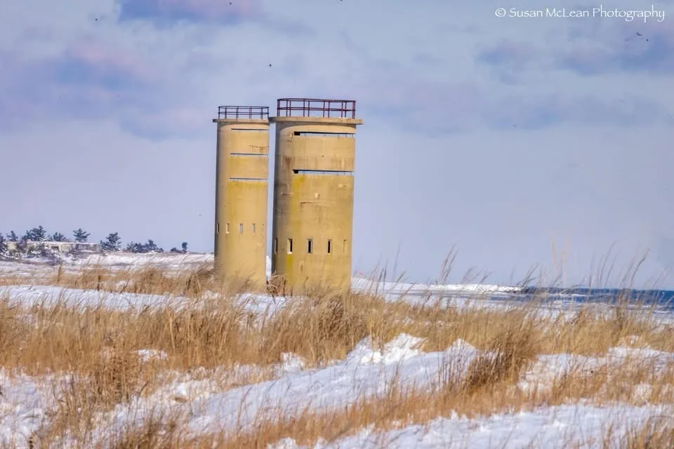 Two weathered concrete towers stand side-by-side in a snowy, windswept coastal landscape with dry grass and a distant ocean…
