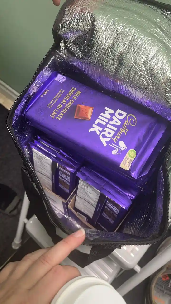 A person holds open a black insulated bag revealing multiple purple-wrapped Cadbury Dairy Milk chocolate bars, with a finger…